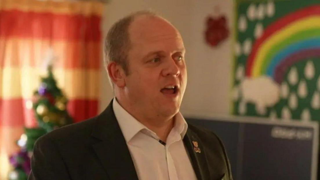 Gareth Pierce is looking away from the camera and is talking. He is wearing a white shirt and dark jacket with a teddy bear pin badge. A Christmas tree and a colourful rainbow mural can be seen in the background of a room at the hospice.