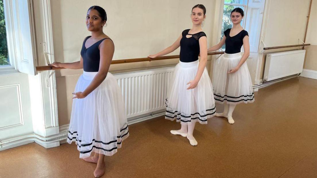 Jemima and two other young female ballet dancers. They are holding on to a bar and they are wearing black leotards and white skirts. Jemima has tights and shoes that match her darker skin tone where as the other paler dancers have white. 