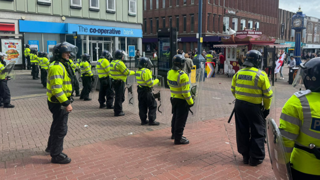 Riot police holding plastic shields lined up along a high street, a group of people is seen in the distance, some of them have St George's flags. Shops are visible in the background