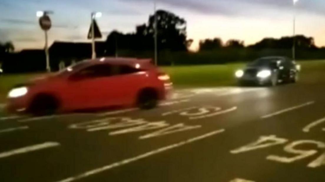 A blurry image showing two cars street racing across multiple lanes on a busy road somewhere in the West Midlands. They are blurry as they are moving a speed. Both have lights on and one is red and one is black. The image has been taken in the early evening or night time. 