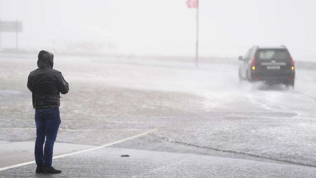 A car driving through floodwater in Galway which a person looks on. 