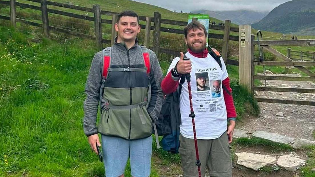 This image shows two men standing with greenery surrounding them, the man on the left is wearing light blue walking trousers and a green and grey jacket, the man on the right is wearing khaki trousers, a long sleeve red top and a white charity t shirt.