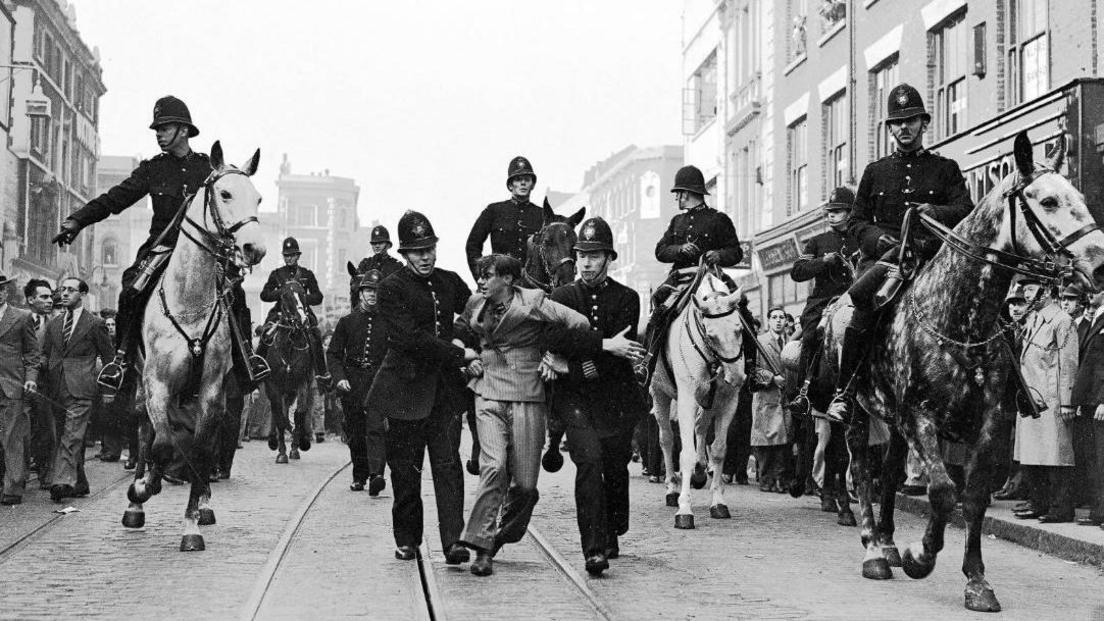 A black and white image of an  anti-fascist demonstrator being taken away under arrest by police after a mounted baton charge during the Battle of Cable Street, London, 4 October 1936 