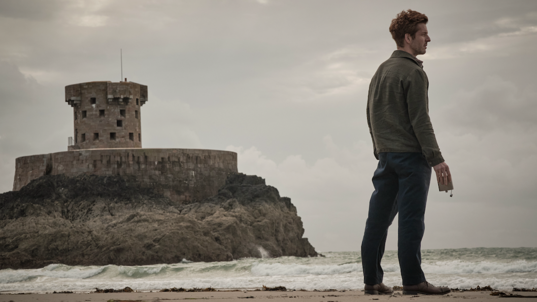 Damien Molony is holding an empty alcohol flask looking out onto the ocean. He's standing on St Ouen's Bay with La Rocco tower and a moody, grey sky behind him.