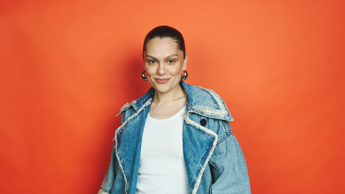 Jessie J is standing against an orange background and smiling at the camera. Her black hair is tied back and she is wearing silver hoop earrings. She is also wearing a white top and jean jacket.