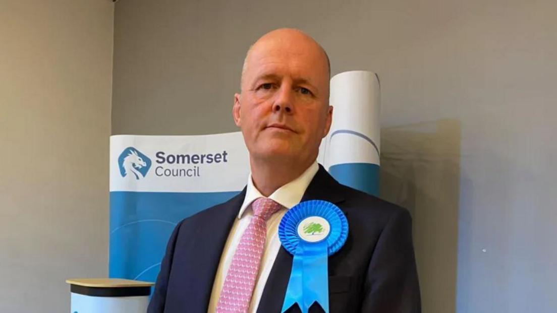 Ashley Fox wearing a suit and pink tie with a blue Conservative badge on. He is standing in front of a Somerset Council banner in a grey-walled room.