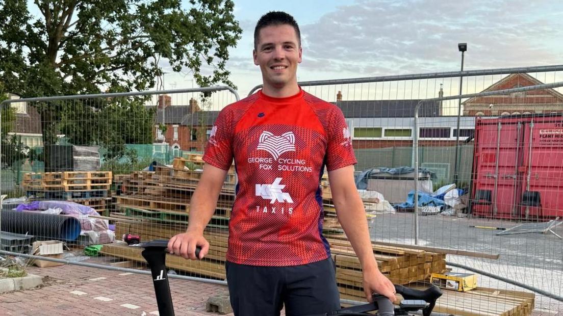 George Fox in a red technical T-shirt with 'George Fox Cycling Solutions" written on the chest. He is holding the saddle and handlebars of a black bike and standing against the backdrop of a construction site.