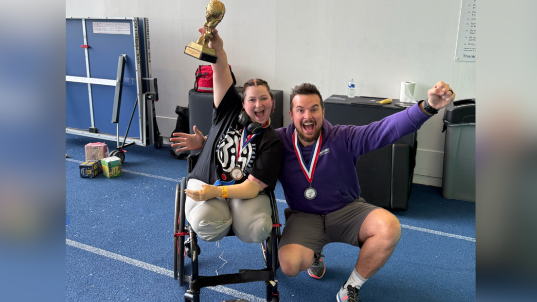Louise Greer is sitting in a wheelchair while lifting her gold trophy. Kneeling next to her is a man with a medal around his neck and a purple hoody. They are in a gym-type room which has a blue floor with white lines painted on it