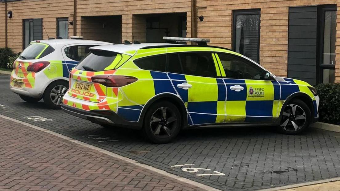 Two police cars are parked outside a modern set of flats, which have cream coloured bricks and windows with black rims.