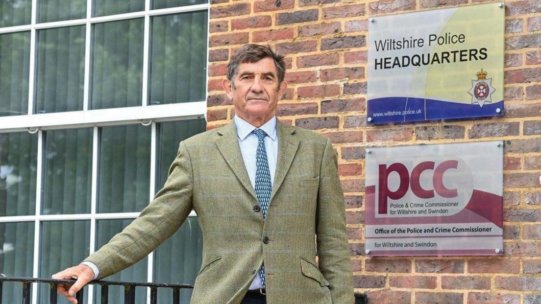 Philip Wilkinson in a white shirt, blue tie and green suit. He stands in front of a brick building which includes a sign for Wiltshire Police and the Police and Crime Commissioner.