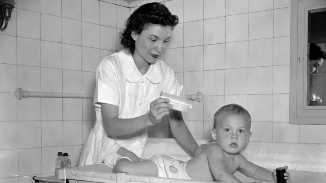 A black and white photograph of a woman, who is dressed in a white dress and holding talcum powder, while a baby in a nappy is laying on their stomach.