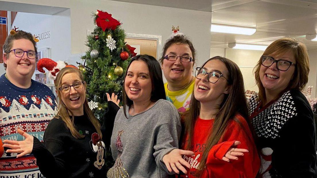 A group of workers from the UK Power Networks call centre in Ipswich are gathered together around a Christmas tree smiling with their hands raised out either side of them. They wear Christmas hats, jumpers and other festive decorations on them.