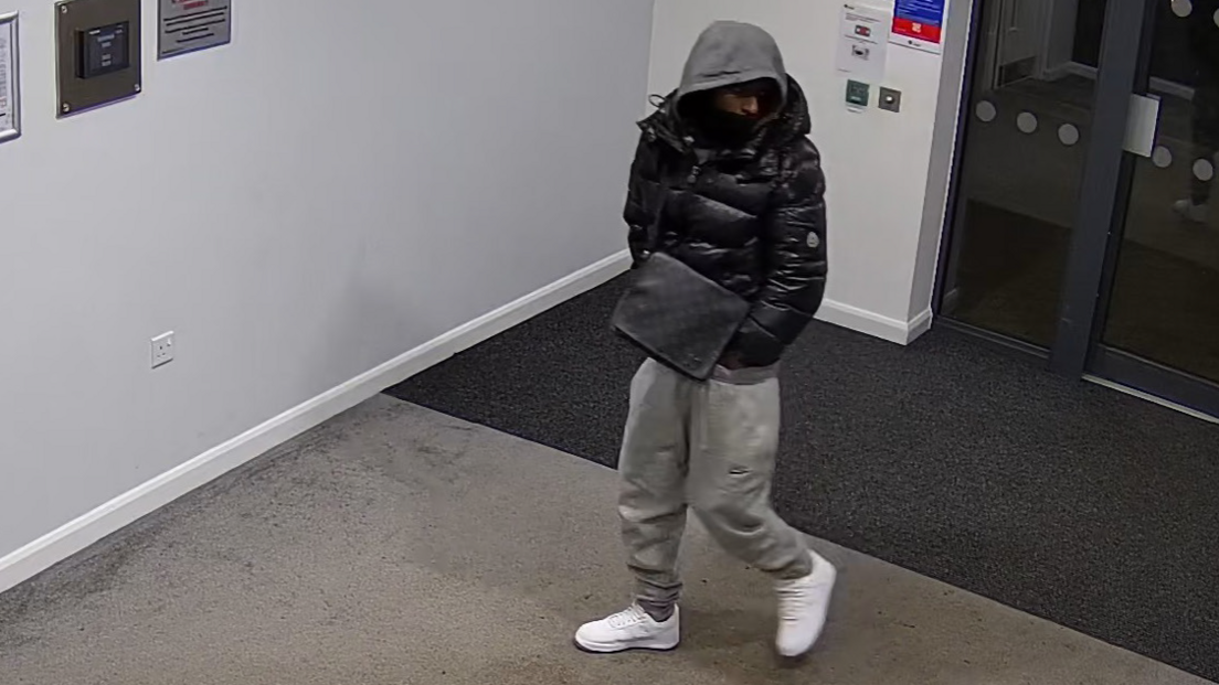 Dualla Abdi walks through the entrance of a building wearing grey tracksuit bottoms, white trainers, a black jacket and his grey hood pulled over his head. The Louis Vuitton bag is black and is worn across his body as a shoulder bag. 