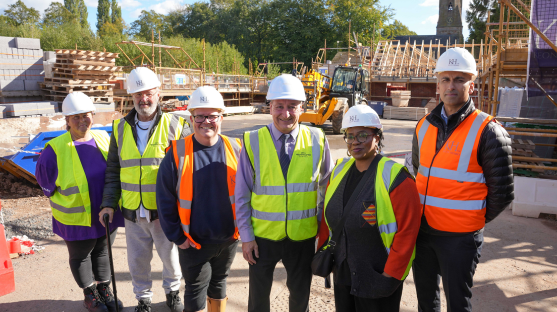 Six people stand in a line, wearing a mixture of yellow and orange hi-vis waistcoats. They are all wearing a white hard hat and standing on a housing development that is work in progress. In the background you can see wooden rafters of roofs being build, with builders and equipment dotted about.

