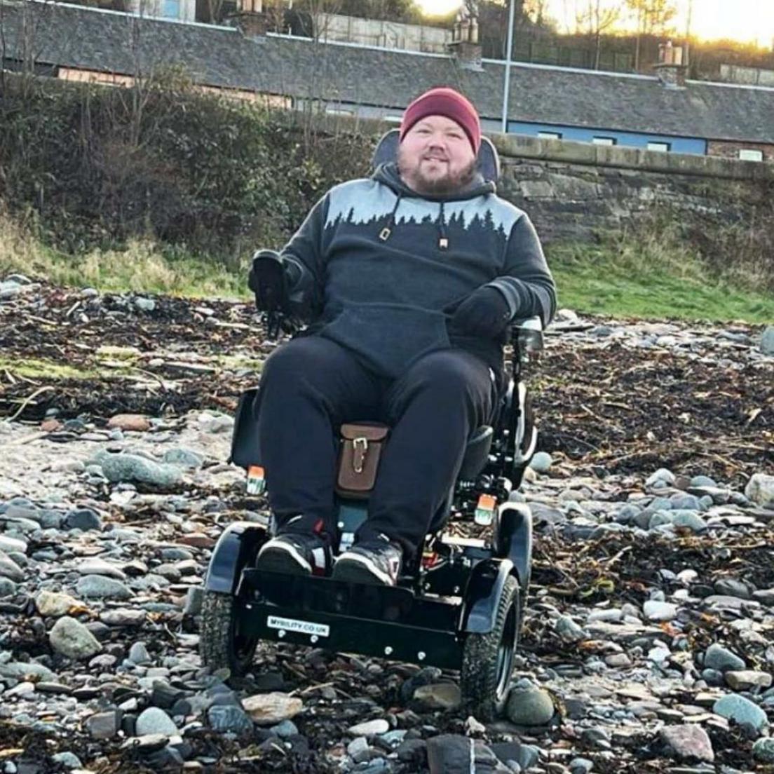 Lucas Honey sitting in a wheelchair on a pebble beach. He is smiling at the camera and is wearing a maroon coloured hat. He is wearing all black clothes and a black hoodie with a grey pattern on the chest.