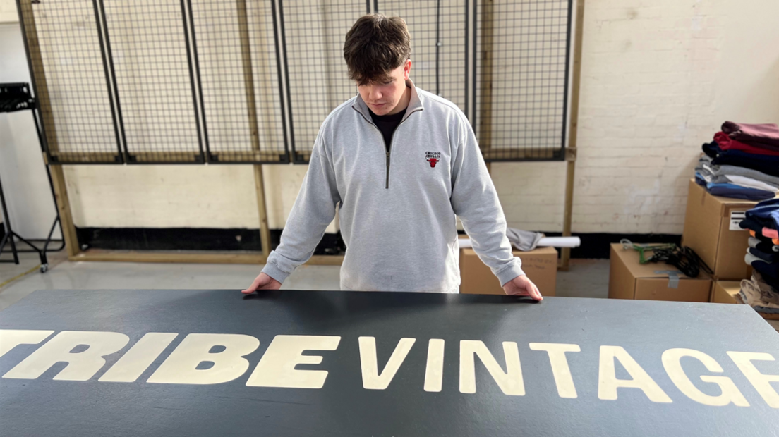 A young man wearing a grey stands behind a sign reading "Tribe Vintage".