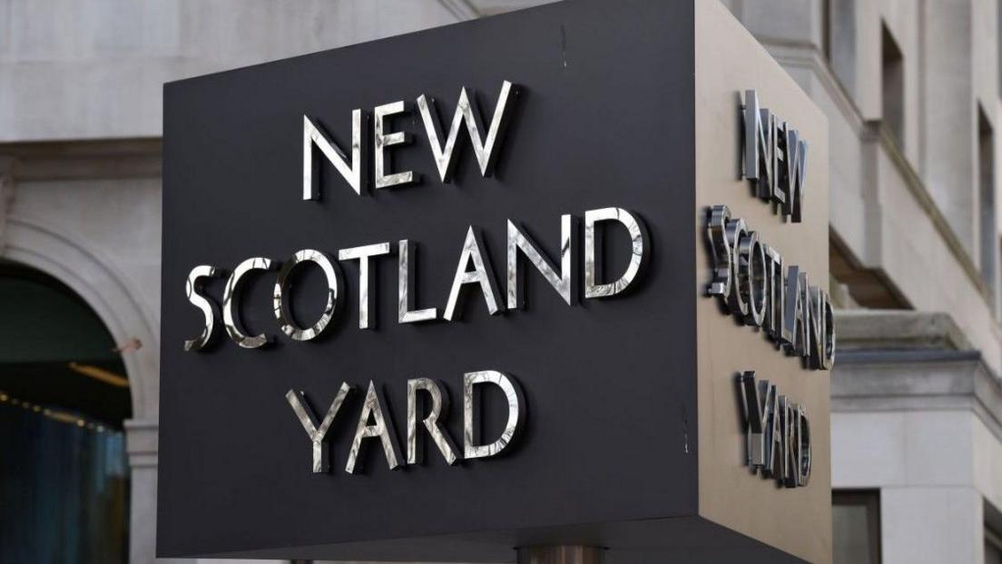 Close-up of a sign which is the shape of a box and says New Scotland Yard on all sides. Pictured outside a building.