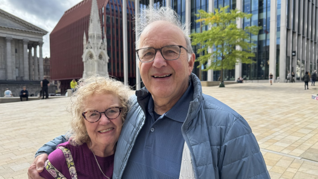 Audrey and Keith Wilkinson in an embrace. They are standing in Birmingham's Chamberlain Square looking at the camera. Audrey is wearing glasses and purple top. Keith is wearing a blue outdoor jacket and a blue polo underneath.