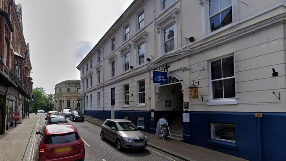 The outside of the Great Malvern Hotel in Graham Road, with pavements on both sides of the road, cars driving down the street and double yellow lines on one side.