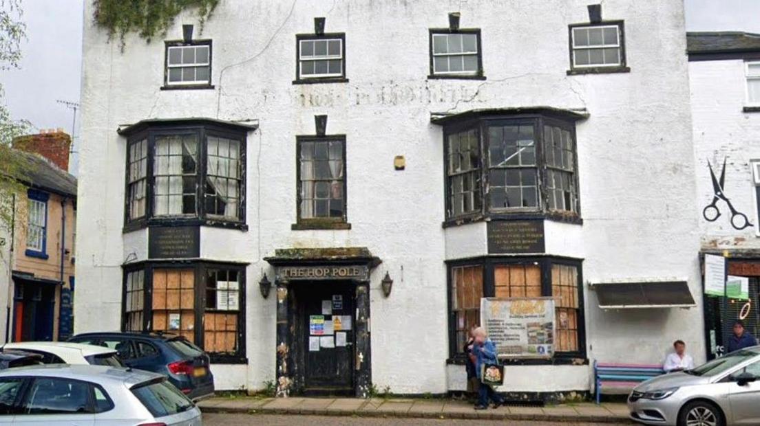 The derelict frontage of a hotel building. There are three storeys, and the building is painted white. There are bay windows on the ground and first floors. There are a number of cars parked in front of the building.