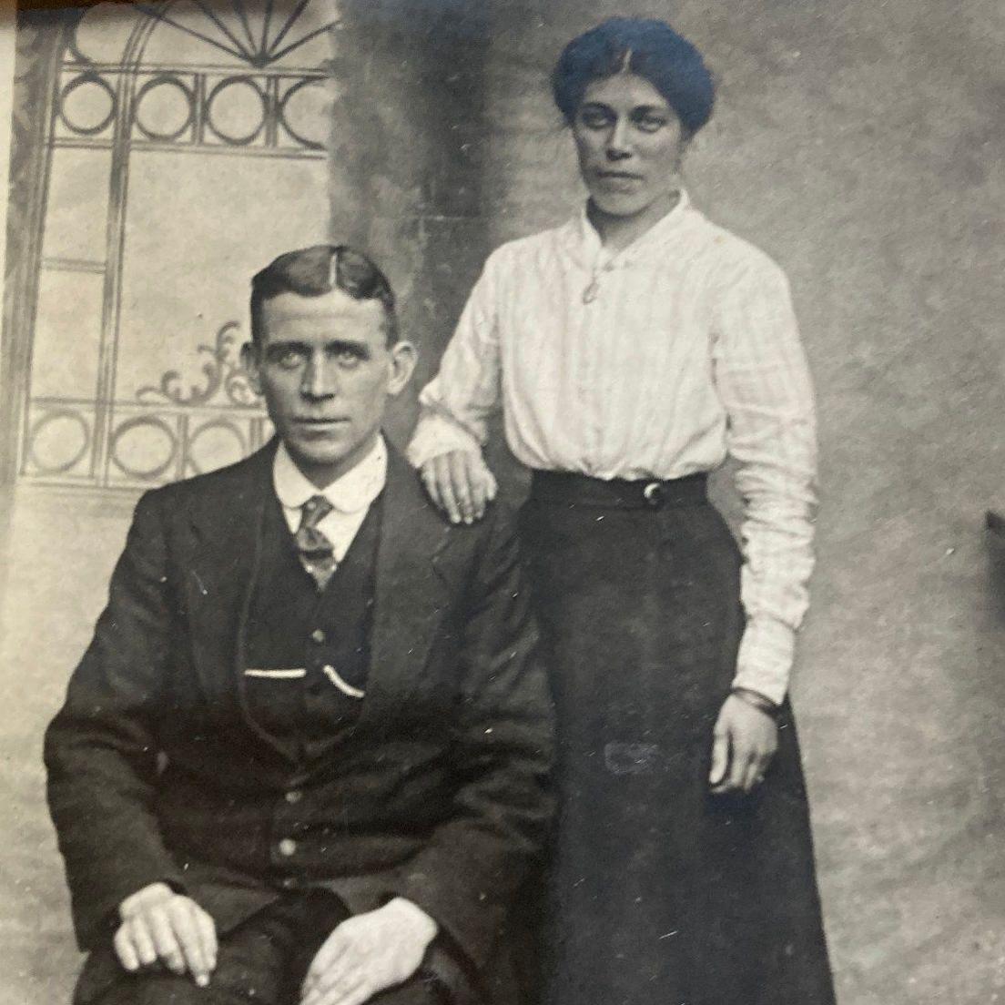 A black and white photograph of man sat in a three-piece suit, with a side parting in his hair, and a woman in a white shirt and long black dress stood behind his shoulder, with her hand resting on him