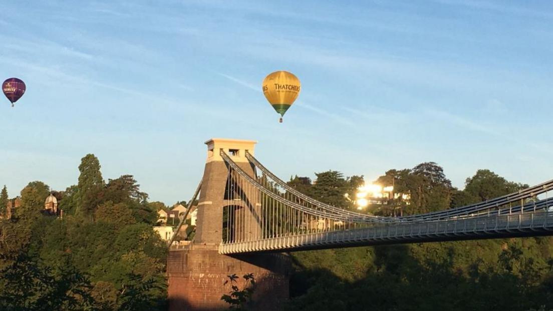 Clifton Suspension Bridge in Bristol. It is a bright sunny day with hot air balloons floating over the bridge. 