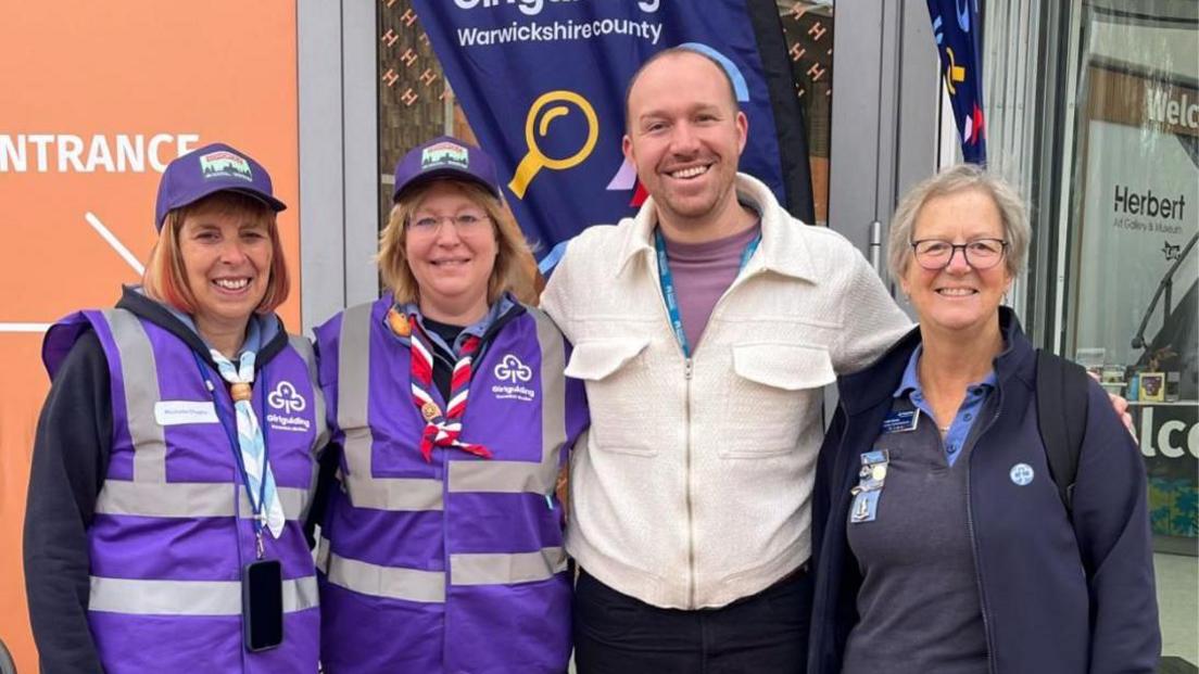 A group of four adults stand in front of a building. There are two women wearing purple vests that have Girlguiding logos. There is also a man wearing a cream cardigan and a woman wearing a dark blue fleece.