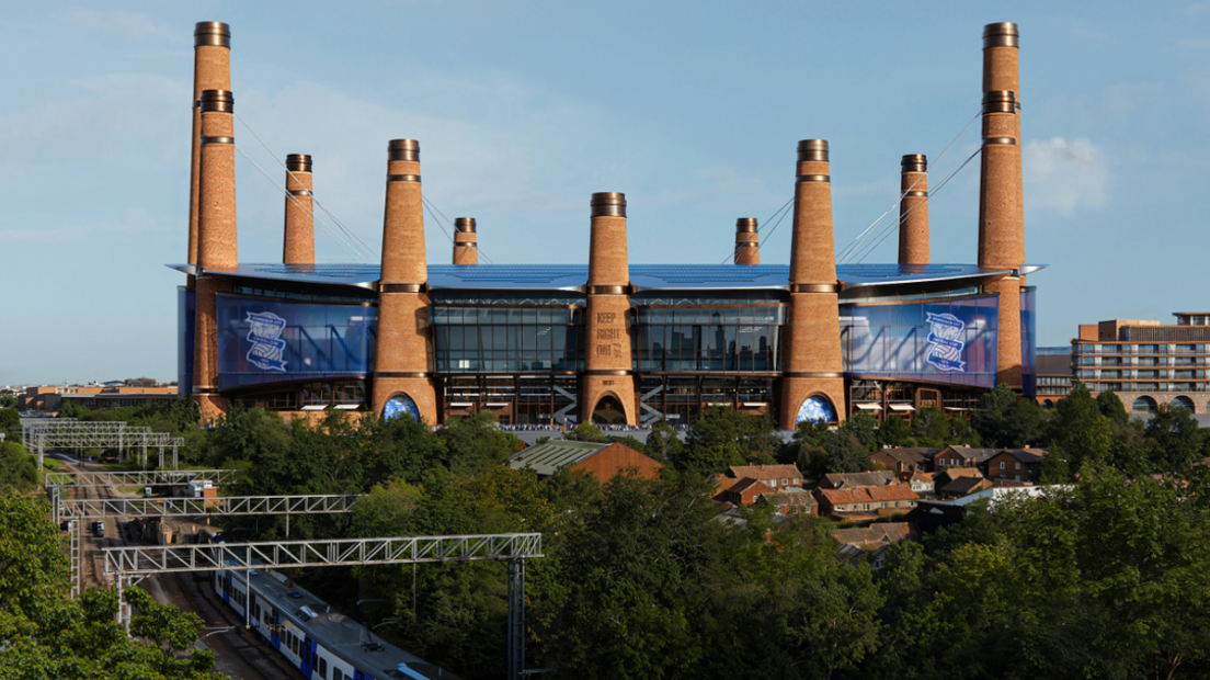 A side view shows almost a dozen chimney towers around the ground which stands behind some housing and railway track to the left.