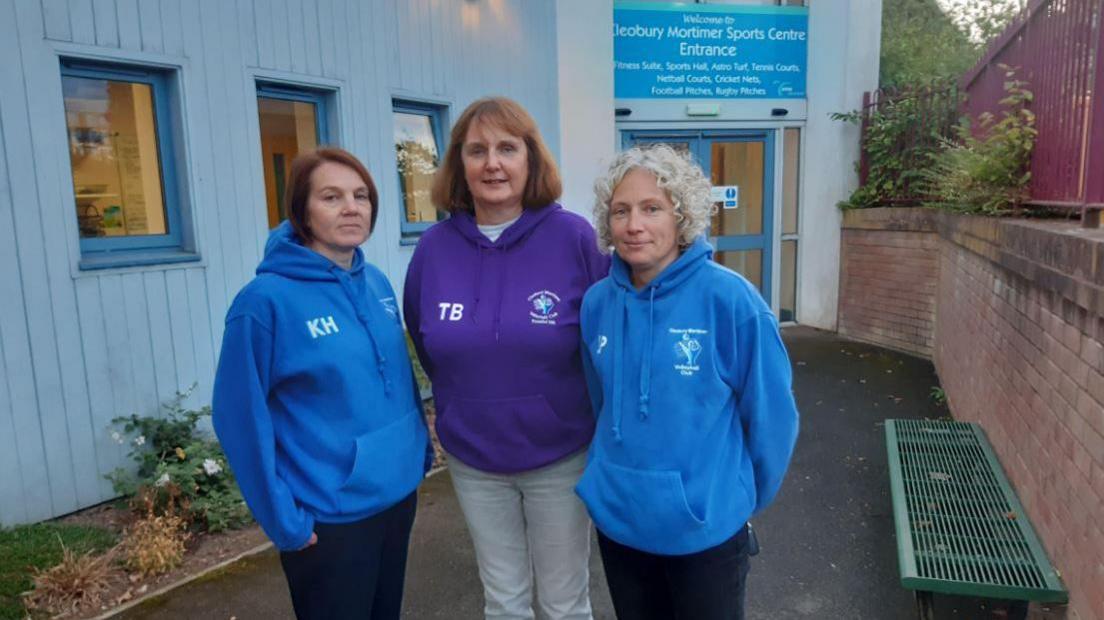Three women standing in front of the entrance to a grey building with a blue sign above the door. One woman has brown hair and is wearing a blue hoodie, a taller woman in the centre has brown hair and is wearing a purple hoodie and the third has blonde hair and a blue hoodie