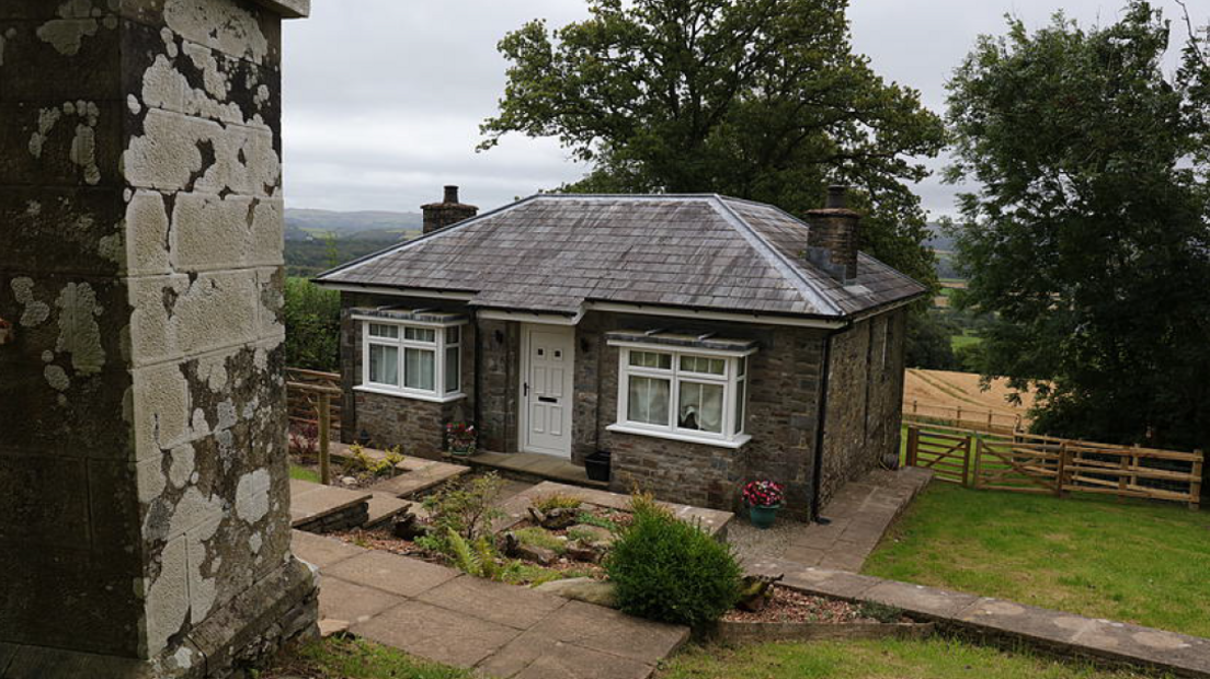 A small grey brick bungalow with white trim windows and a door, with a farmer's field and woods in the background. 