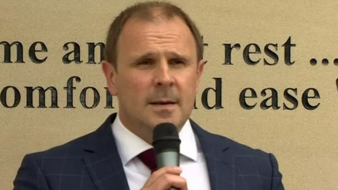 Winston Irvine is wearing a blue checked jacket, white shirt and red tie, and is holding a microphone