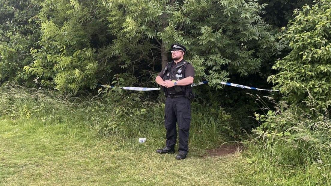 Policeman with police tape at the scene of the incident. Green bushes are around the cordon and a path is visible.