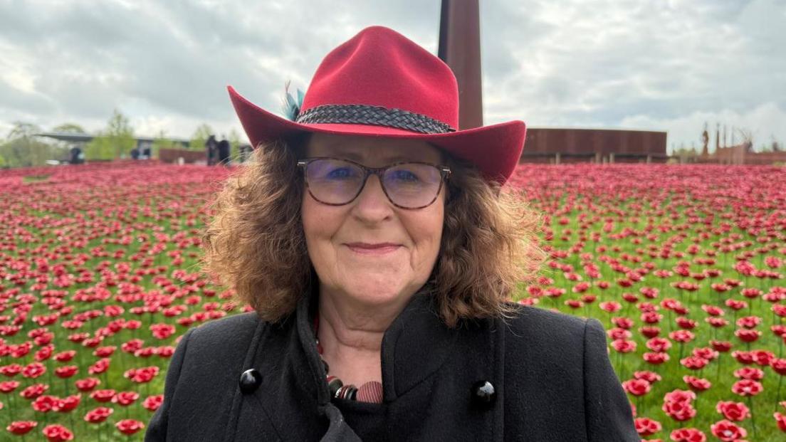 A lady is smiling at the camera. She has brown hair and a red hat and is wearing glasses. Behind her are thousands of ceramic poppies planted in the ground.