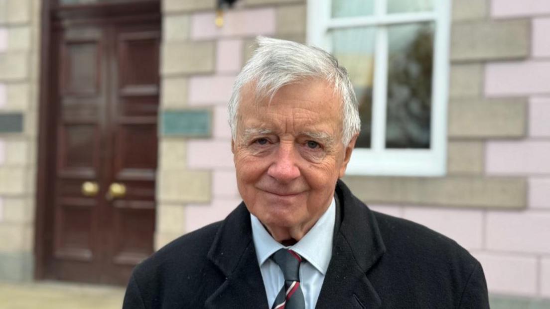 Philip Bailhache smiles for the camera. He is stood outside the states building in the Royal square. He is wearing a black wool coat, blue shirt and stripy red and green tie. He has white hair. 