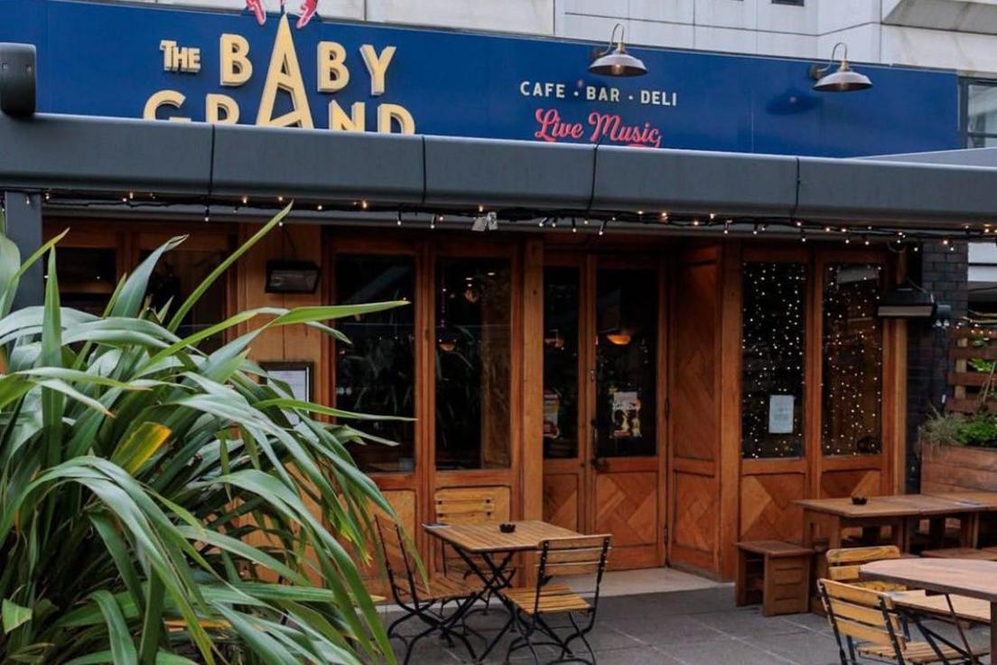 An outside view of the Baby Grand piano bar. There is a plant in the foreground. There are tables and chairs outside. A sign above the door, which is surrounded by wood, is blue, with the name of the bar in yellow. Another sign, reading 'Elmbank Gardens' is up high in the background on a light-coloured building.