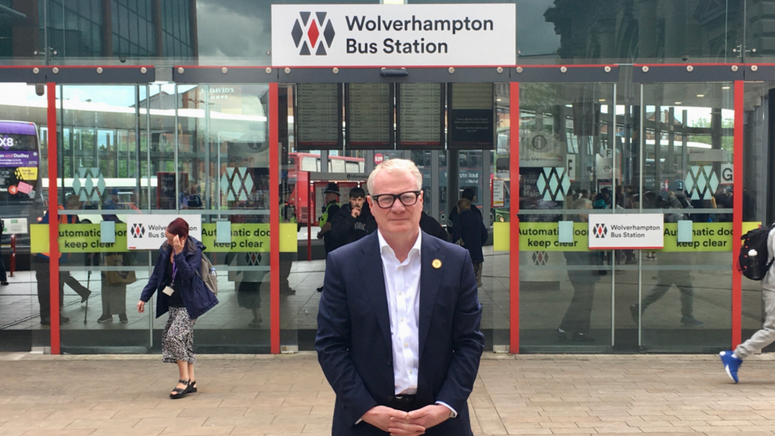 A man with short white hair and glasses is wearing a navy blue suit jacket over a white shirt with an open collar. He is standing with his hands clasped in front of a set of glass doors under a sign reading "Wolverhampton Bus Station".