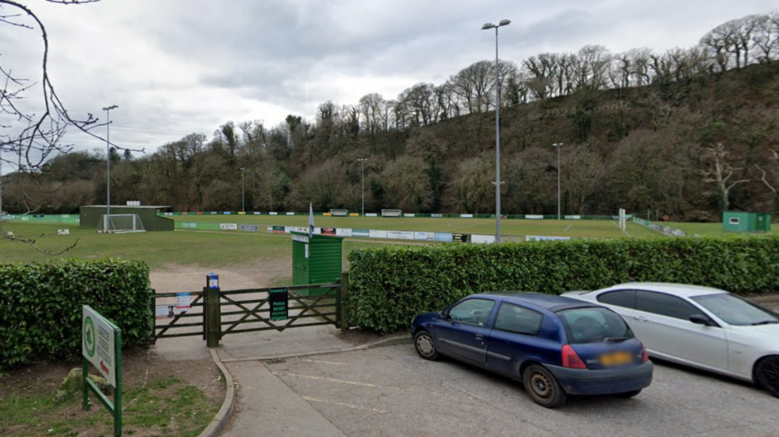 Ivybridge Town Football Club. Cars are parked outside the football club. A hedge separates the car park from the pitch. The green pitch is surrounded by trees.