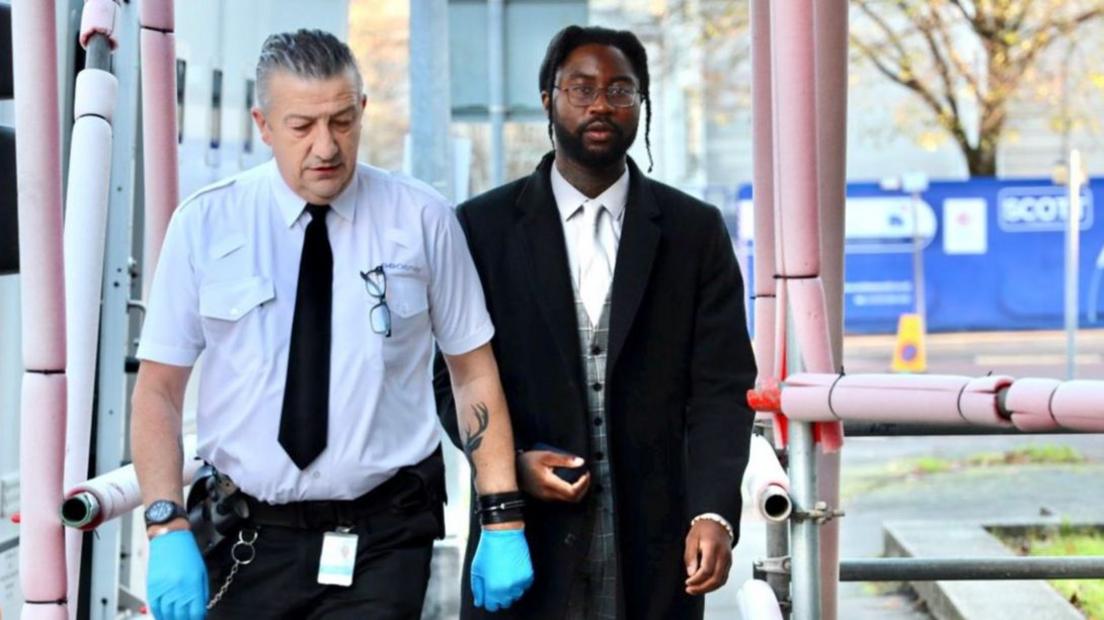 Joshua Gordon arriving at court wearing glasses, a black jacket and a suit. He is accompanied by a prison warden with a white shirt and black tie. The warden wears blue gloves.