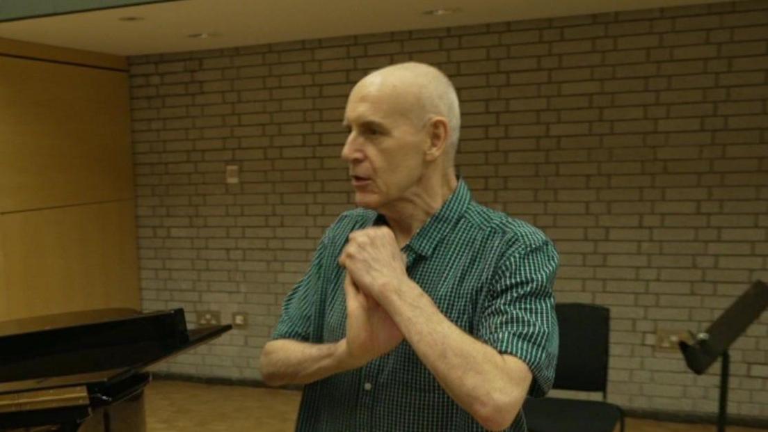 Mick Broad clasps his hand together wearing a green-striped shirt, smiling as he listens to a performance on a piano inside an auditorium. 