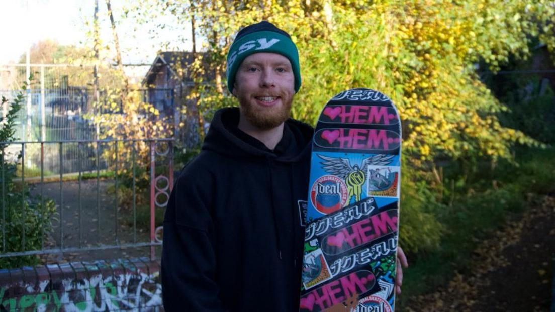 Shaun Boyle is wearing a green and black woolly hat and a black hoodie. He is holding a colourful skateboard filled with different patterns and graffiti art