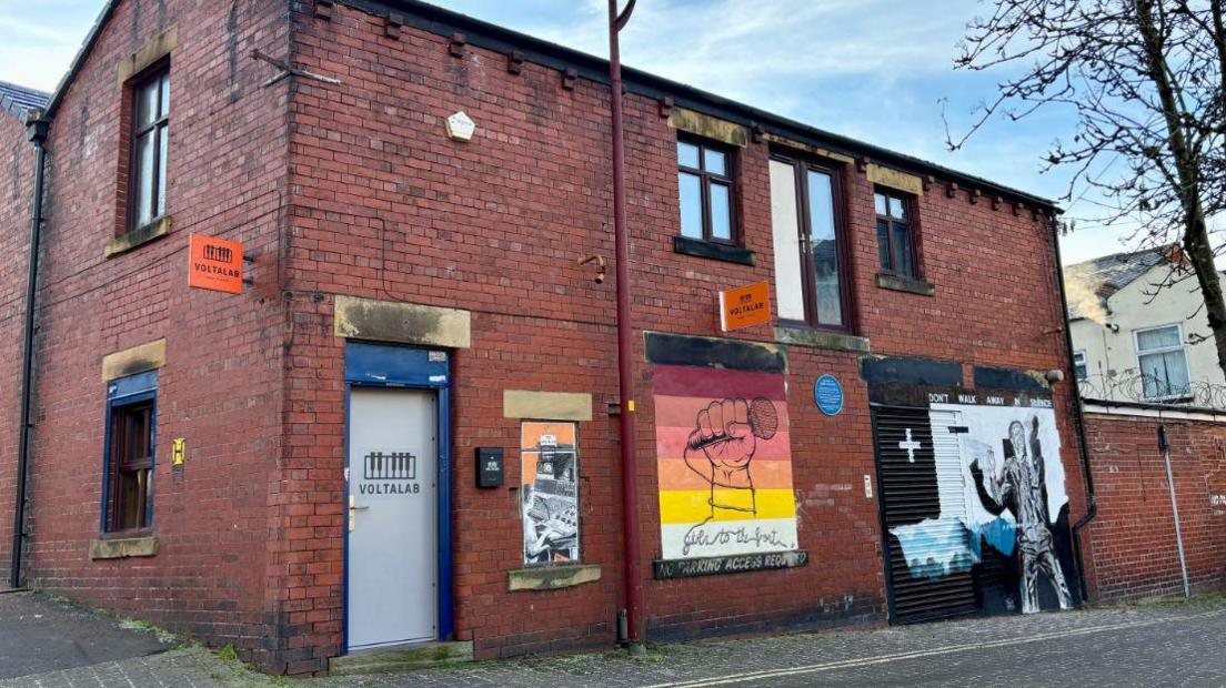 The redbrick Voltalab building with band posters and the Curtis mural seen from the roadside on a clear day. 