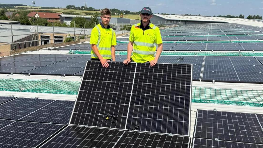 Two men in yellow high-vis jackets stand among dozens of solar panels on the roof of a building