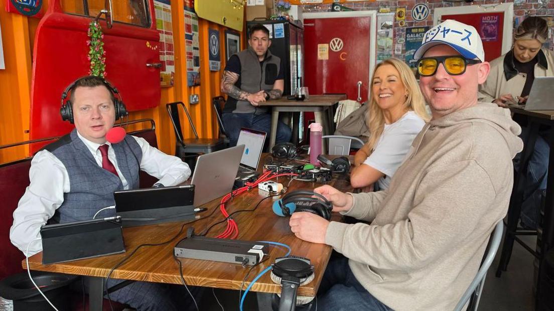 Matt Chorley, in a waistcoat and tie, speaks into  red microphone in a Gateshead cafe, across the table from listeners Joanne an Ben, who are both grinning broadly
