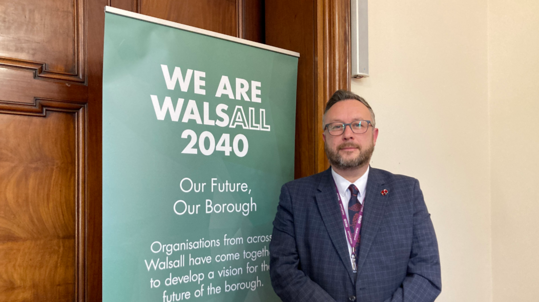 Pictured is Garry Perry with a slight smile on his face. He is wearing round dark framed glasses, has a dark blond quiff and a dark blonde beard. He is wearing a dark blue blazer with a white shirt underneath and a blue and red tie. He is standing next to a green sign that says "We are Walsall 2040".