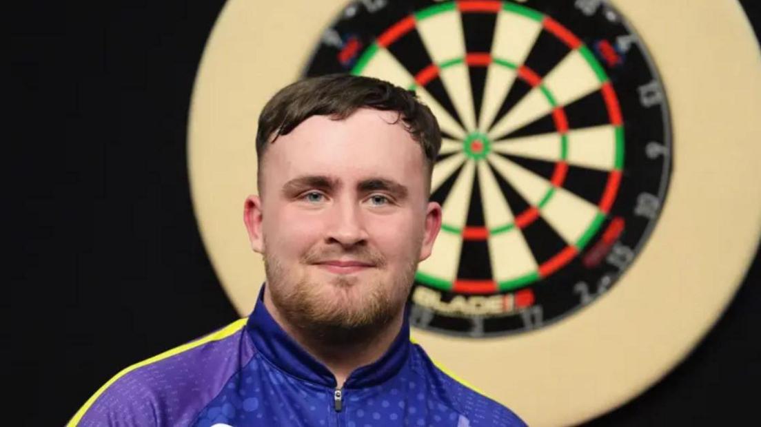 Head and shoulders shot of dart player Luke Littler wearing a purple zip-up sports shirt, with a dart board in the background