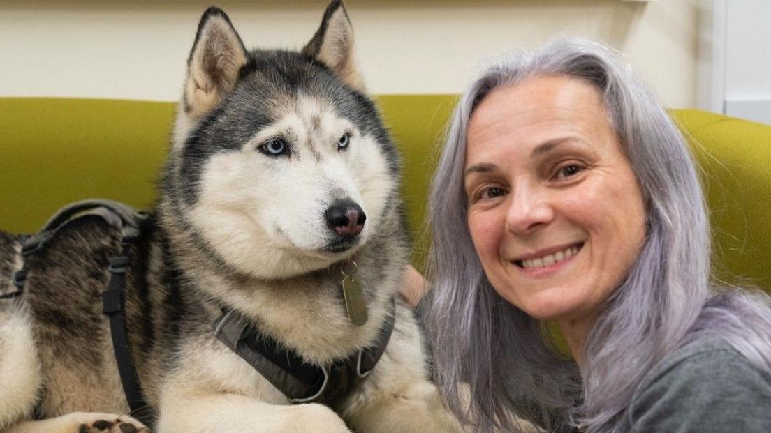 A grey-and-white, blue-eyed husky dog lies down on a mustard-yellow sofa. She is wearing a harness and a collar with a metal tag. To her right is Prof Clara Mancini, who has long, lilac hair and is wearing a grey top. She is smiling at the camera.