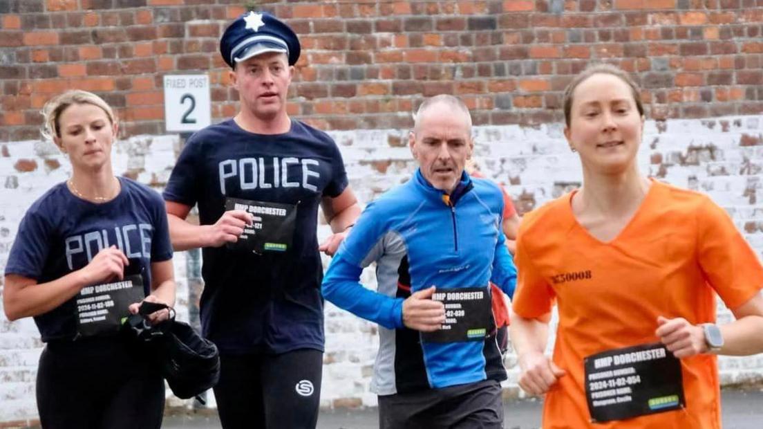 Four runners, two men and two women, running towards the camera in Dorchester prison yard. One man and woman are dressed as police officers, the other woman is dressed in an orange prison uniform. The other man is dressed in shorts and a long-sleeve running top.