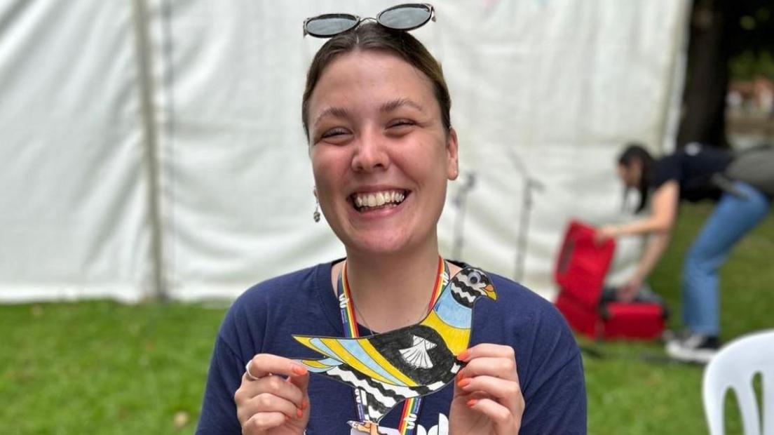 A woman with sunglasses on her head and wearing a navy T-shirt holds her black, blue, yellow and white design of a coloured-in pigeon. 