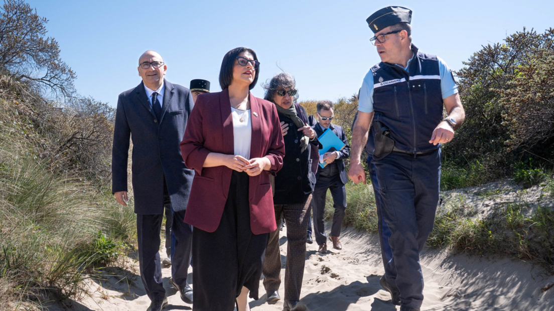 Home Secretary Shabana Mahmood is wearing a black skirt, a white top and a maroon jacket. She is walking along a sandy path next to a French law enforcement officer. There are four people walking behind her including French Interior Minister, Laurent Nunez who is wearing a suit.