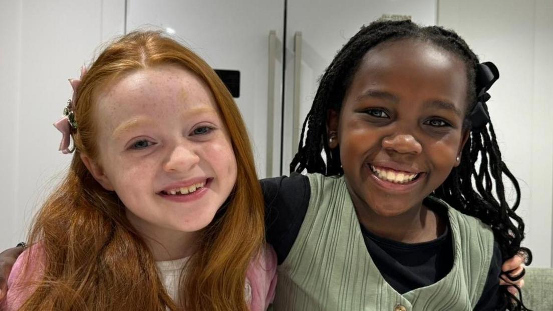 Two girls are hugging whilst smiling at the camera. One girl is wearing pink and the other girl is wearing green. 